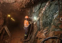 Fresh Investment Cycle in Gold Mining Sector in Zimbabwe Mine worker in orange safety gear drilling into rock inside a dim tunnel with metal mesh overhead and lights in the distance.
