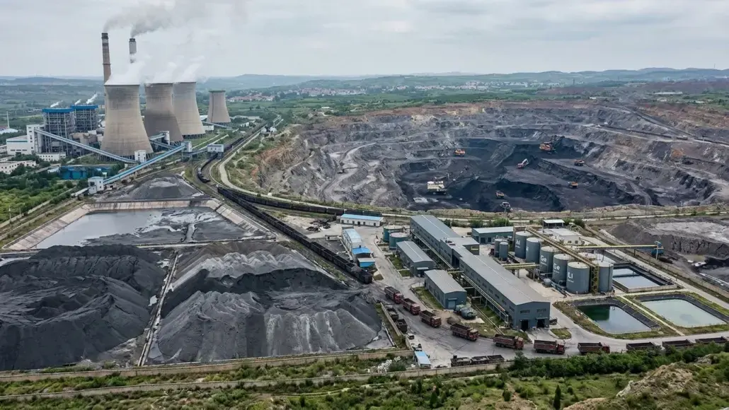 Aerial view of a large industrial complex with cooling towers emitting steam, coal stockpiles, and processing buildings beside rail lines and conveyors.