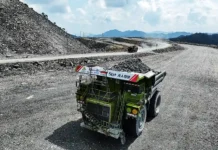 China, Australia Expand Ties on Entire Mining Value Chain Green mining dump truck on a dusty gravel road at a quarry with rock piles and distant hills behind it.