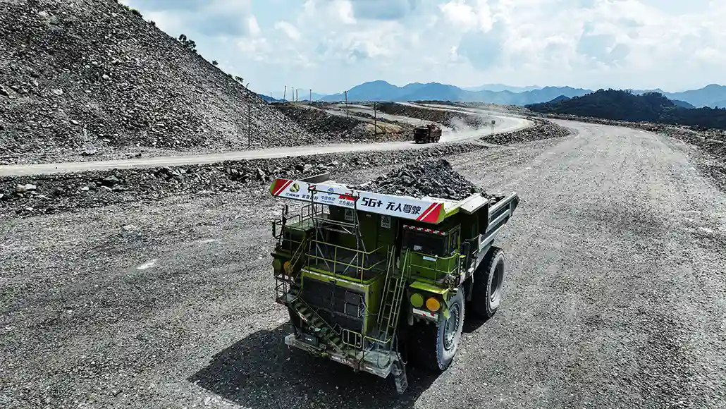 Green mining dump truck on a dusty gravel road at a quarry with rock piles and distant hills behind it.