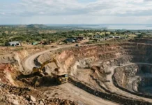 Imwelo Gold Project, A Tanzania Gold Mine to Be Gold Funded Open-pit quarry with a yellow excavator loading rocks into a dump truck, terraced walls and dusty roads in a rural landscape.
