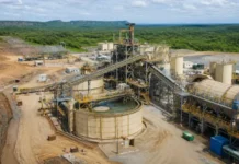 Fortuna Approves Séguéla Gold Mine Expansion Aerial view of a mining processing plant with conveyors, tanks, and metal framework in a dusty quarry surrounded by green hills.