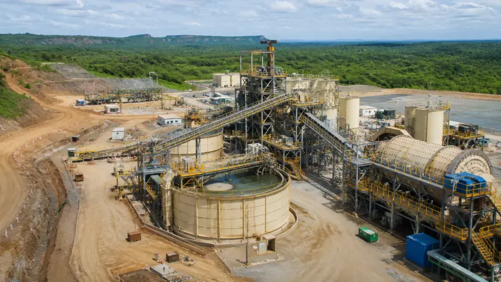 Aerial view of a mining processing plant with conveyors, tanks, and metal framework in a dusty quarry surrounded by green hills.