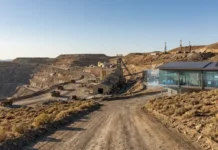 Tonopah West Advances Under US Critical Minerals Strategy Open-pit mine with heavy machinery and a glass-fronted processing building along a dusty road, hillside in the background with clear sky.