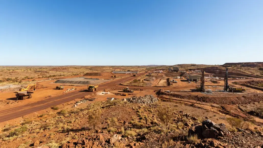 Arid construction site with heavy machinery and drill rigs spread across red dirt landscape under a clear blue sky.