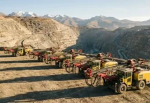 Epiroc Wins Large Order for Mining Equipment in Peru Row of yellow mining trucks with red drill arms lined up along the edge of an open-pit quarry, with rugged canyon walls and distant snow-capped mountains in the background.