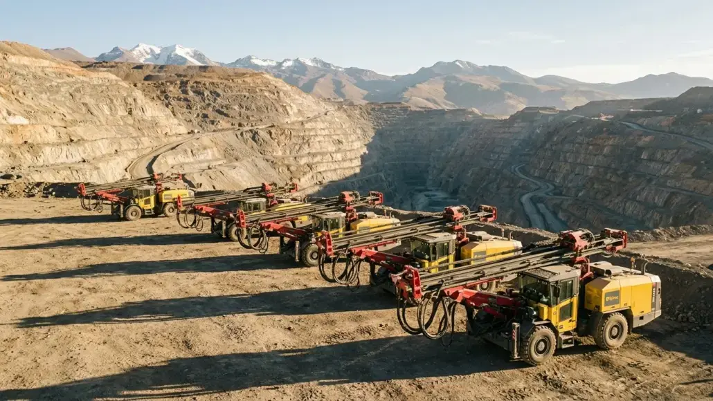 Row of yellow mining trucks with red drill arms lined up along the edge of an open-pit quarry, with rugged canyon walls and distant snow-capped mountains in the background.