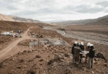 First Quantum Explores Taca Taca Copper Project Stake Sale Desert mining site with temporary cabins and trucks; workers in hard hats study plans on a rocky hillside.