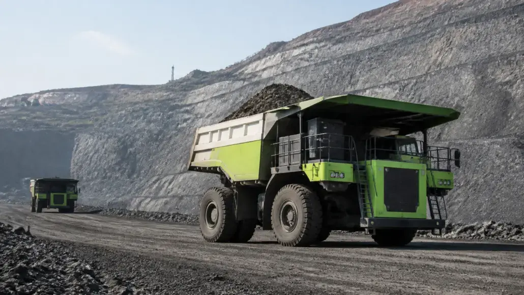 Large green mining dump truck at a rock quarry, on a dusty road with steep rock walls behind it.