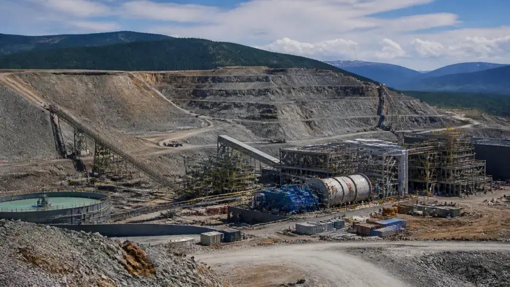 Open-pit mining site with conveyor belts, large machinery, and storage tanks against terraced rock faces and distant hills.