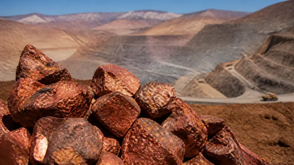 Close-up of reddish brown rocks in the foreground with a desert canyon and winding road in the distance.