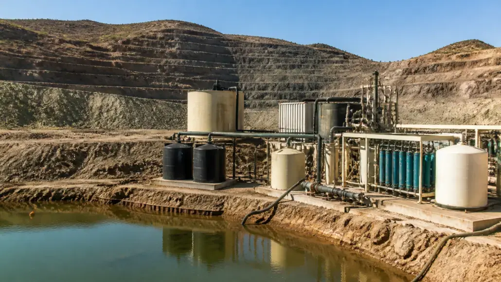 Industrial water treatment setup with pumps, tanks and blue filter columns beside a pond in a rocky quarry setting
