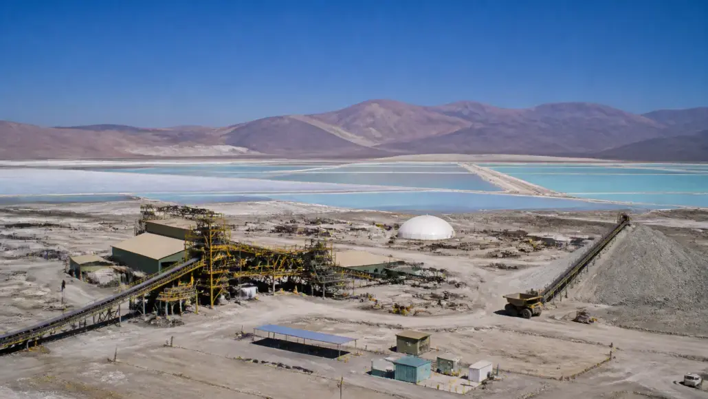Aerial view of a mining facility with yellow scaffolding and conveyor belts beside turquoise salt ponds and distant purple mountains under a clear blue sky.