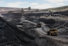Global Coal Mine Pipeline Expands as China and India Lead Open-pit mine with two large dump trucks driving along a dirt road and a conveyor belt system in the background under a partly cloudy sky.