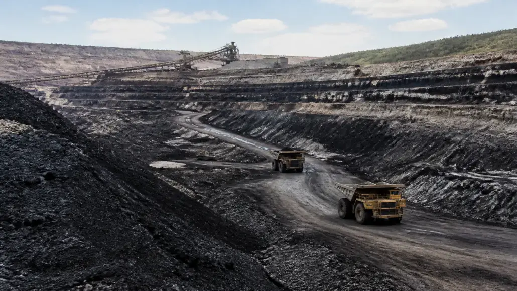 Open-pit mine with two large dump trucks driving along a dirt road and a conveyor belt system in the background under a partly cloudy sky.