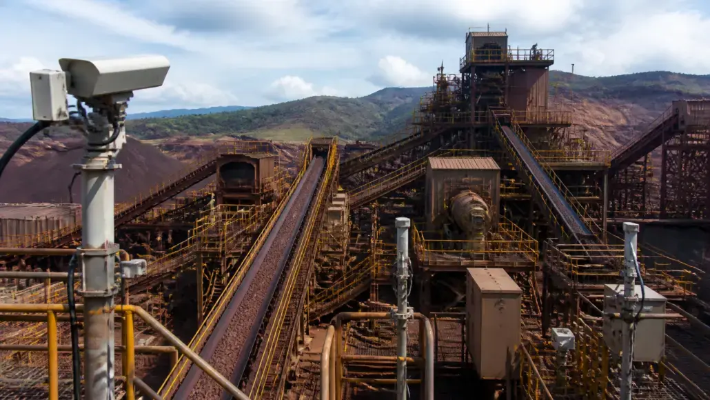 Industrial mining facility with elevated conveyor belts and rust-colored metal structures against a hilly backdrop.
