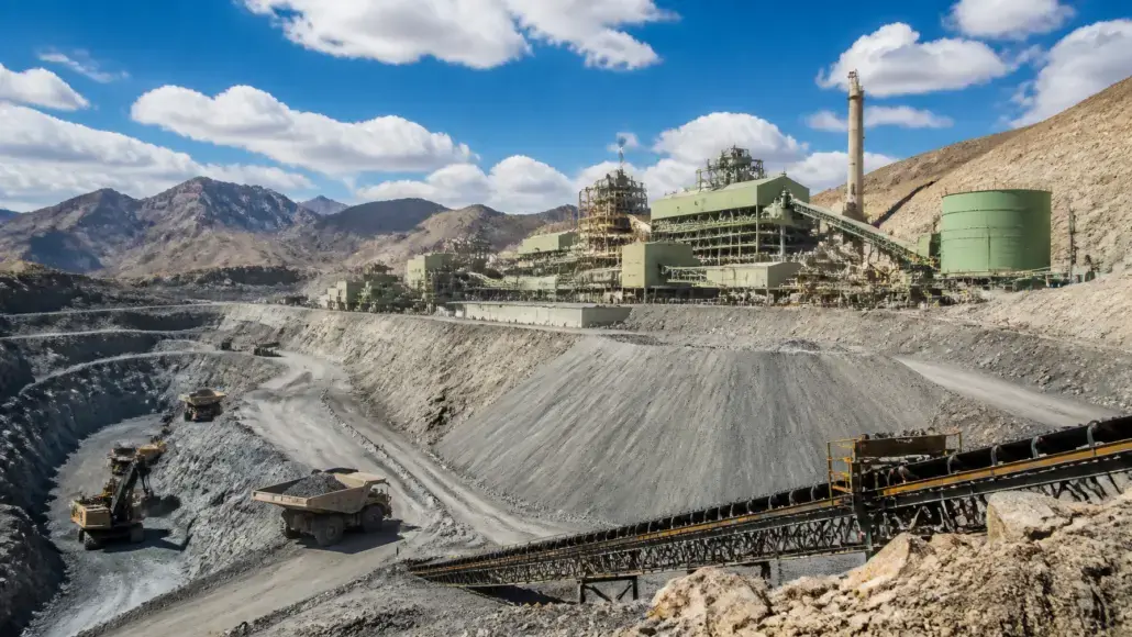 Open-pit mining site with terraced slopes, heavy trucks, and a green processing plant connected by conveyors under a blue sky.