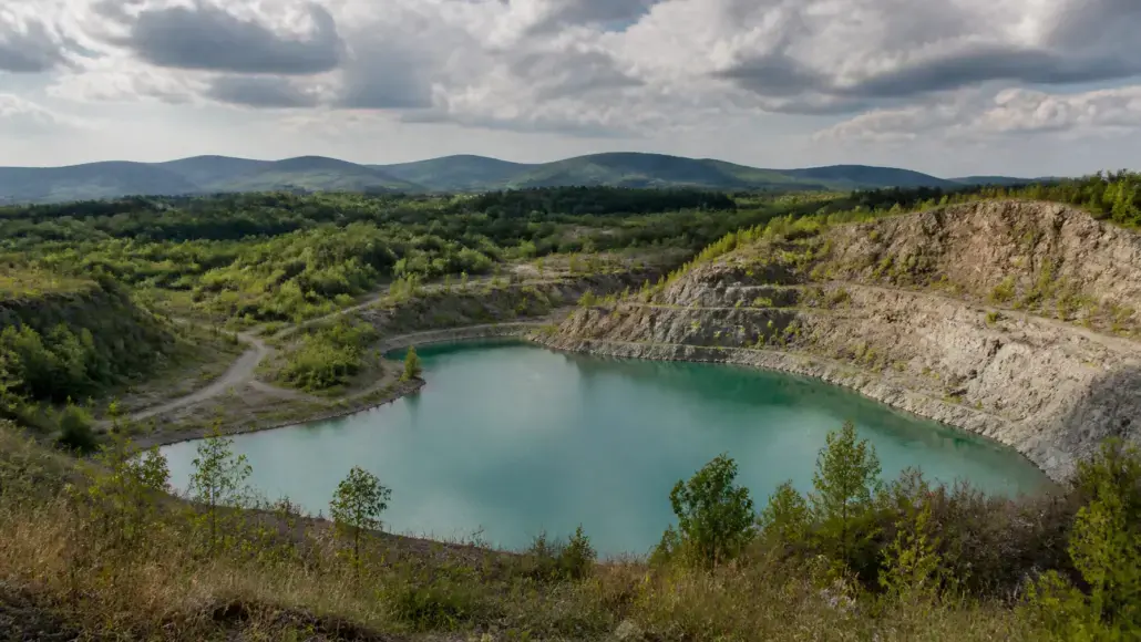 Turquoise pond in a rocky quarry crater with vegetation on slopes and distant hills under a cloudy sky.