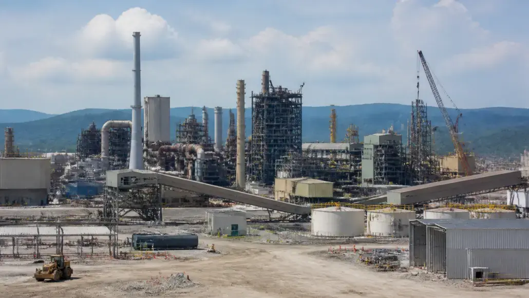 Industrial refinery complex with tall distillation towers, interconnected pipes, and a crane in the background; foreground shows a construction area with equipment.