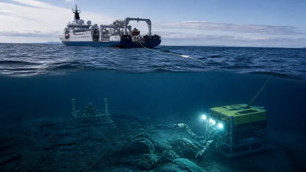 Remotely operated underwater vehicle with lights on the seafloor, tethered to a research ship above.