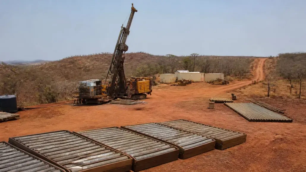 Oil drilling rig on a dusty red dirt site with stacked pipes and equipment nearby.