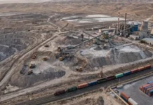 Uzbekistan Invites Indian Companies to Explore Mining Investment and Rare Earth Opportunities Aerial view of a large open-pit mine with conveyor belts, processing buildings, and cooling towers; a colorful freight train runs along tracks in the foreground.