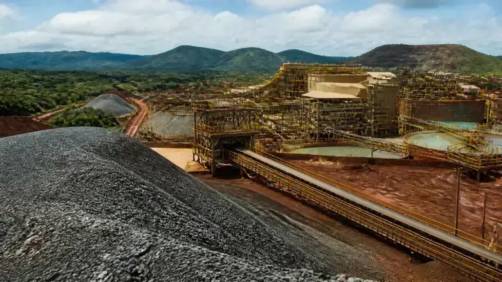 Industrial coal mining site with conveyor belts and large coal piles against a hilly landscape in the background.
