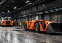 Nornickel and BELAZ Agree to Establish Mining Equipment Joint Venture Underground mining tunnel with orange heavy equipment, including a large front-end loader at the front and a dump truck behind it.