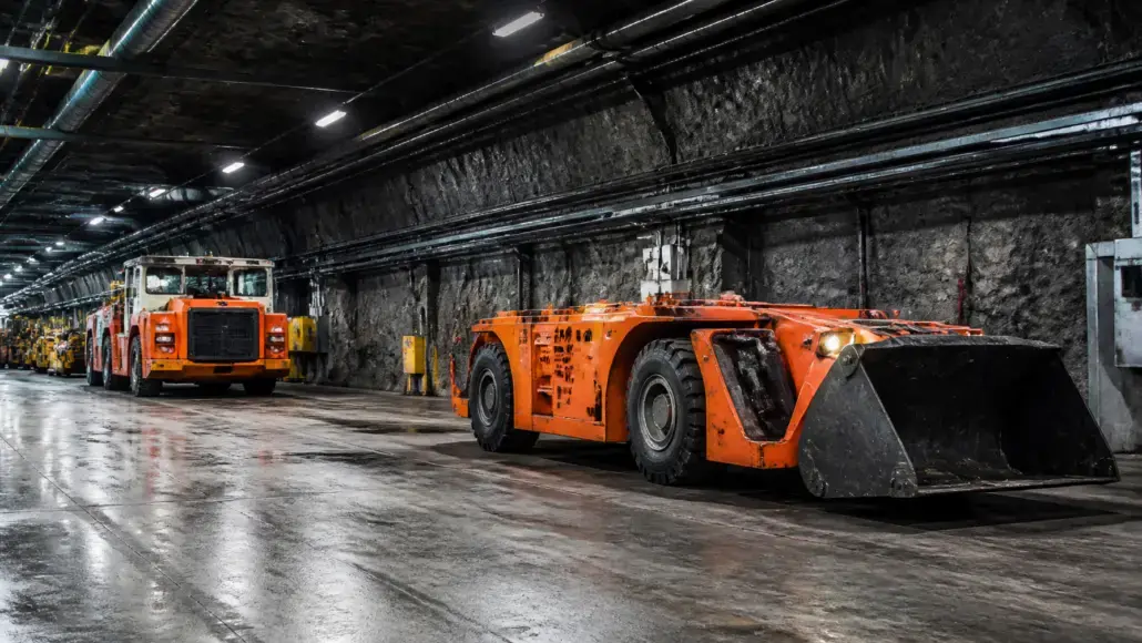 Underground mining tunnel with orange heavy equipment, including a large front-end loader at the front and a dump truck behind it.