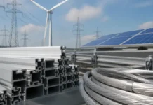 Renewable Energy Expansion Driving New Aluminium Demand Stacks of aluminum structural beams and coiled steel cables at a renewable energy site with solar panels and a wind turbine in the background, under a blue sky.