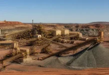 BHP Awards Contracts for South Australia Copper Projects Open-pit mining site with multiple conveyor belts and yellow metal structures feeding large piles of gravel/ore. Red earth surrounds the facility under a clear blue sky.