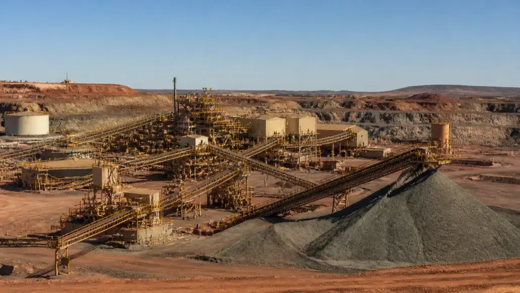 Open-pit mining site with multiple conveyor belts and yellow metal structures feeding large piles of gravel/ore. Red earth surrounds the facility under a clear blue sky.